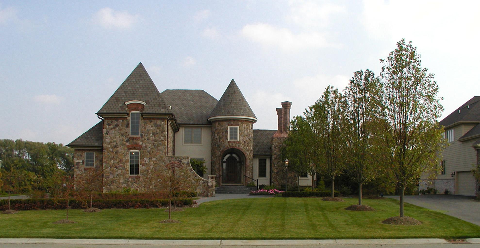 Castle-like stone house with an arched wooden front door, round turret, and landscaped entrance with flowers and trees.