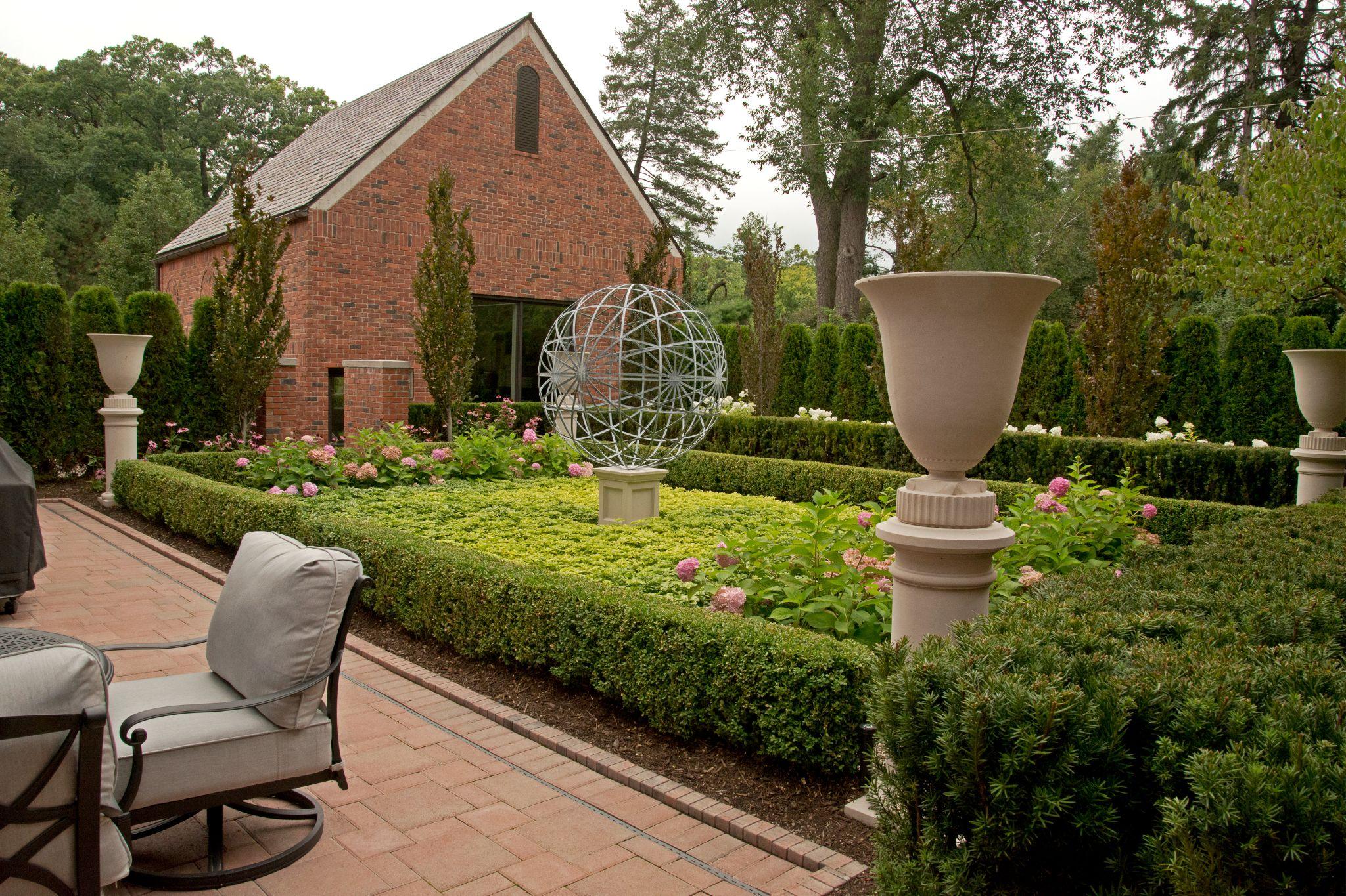 Elegant courtyard garden with boxwood hedges, pink flowers, and a modern metal globe sculpture centerpiece, bordered by large urn planters and a red brick building in the background.