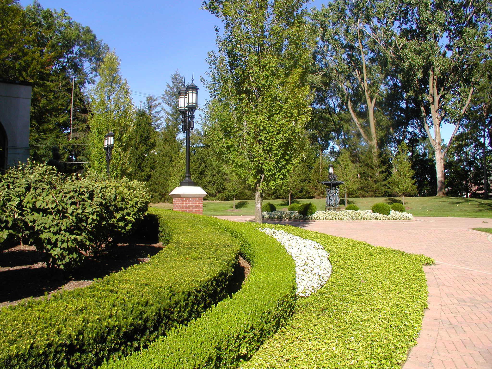 Formal landscaped garden with neatly trimmed hedges in curved patterns, a brick-paved walkway, classic black lantern-style lampposts, and a small fountain in the distance surrounded by trees.