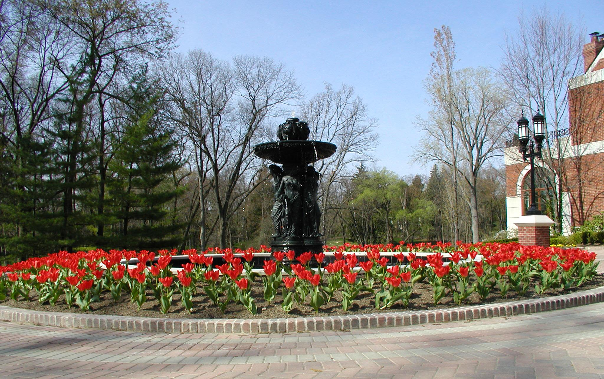 Large red-brick mansion with a circular driveway, central fountain surrounded by flowers.