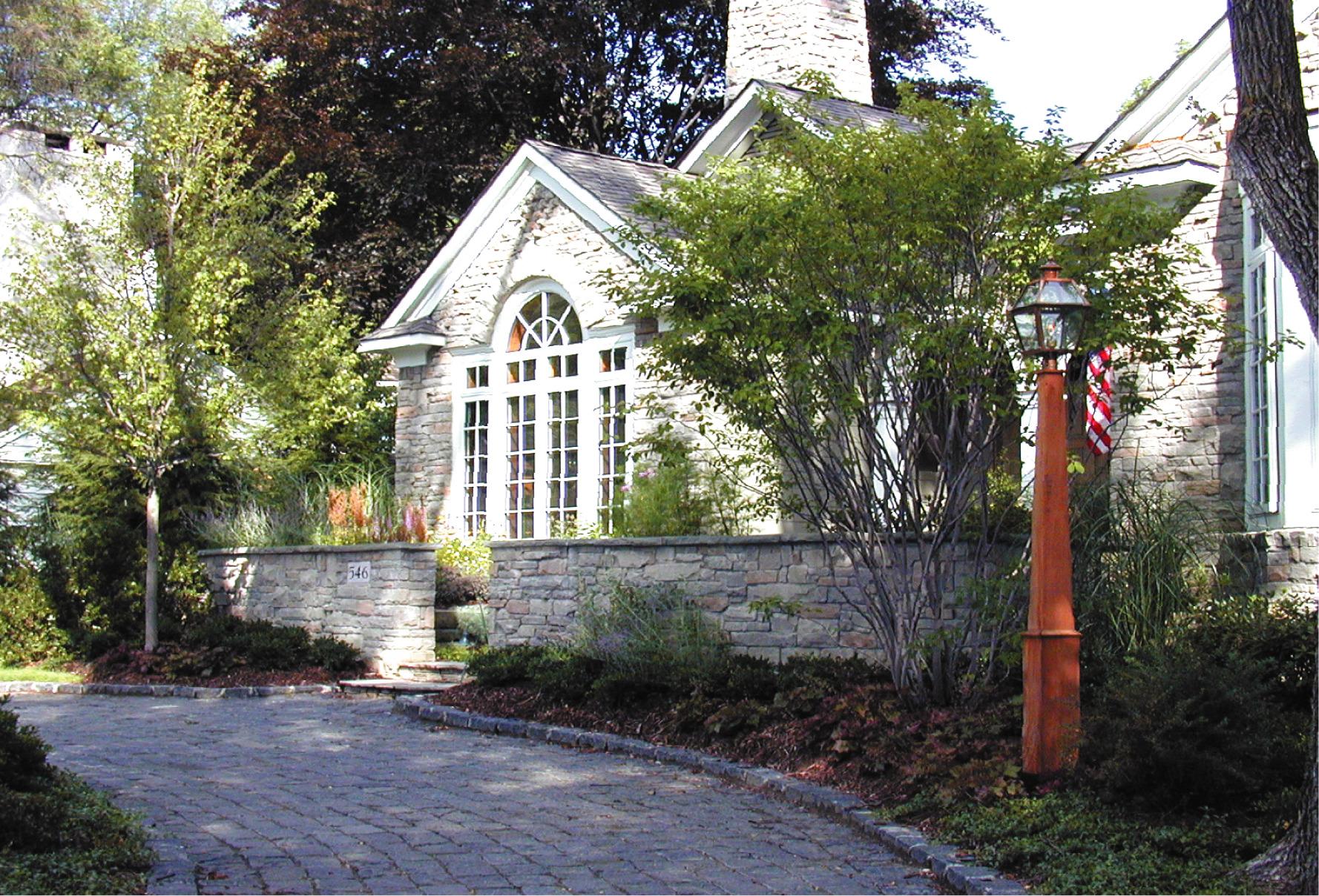Stone house with tall arched windows, surrounded by trees, shrubs, and a stone wall, with an American flag visible near the entrance.