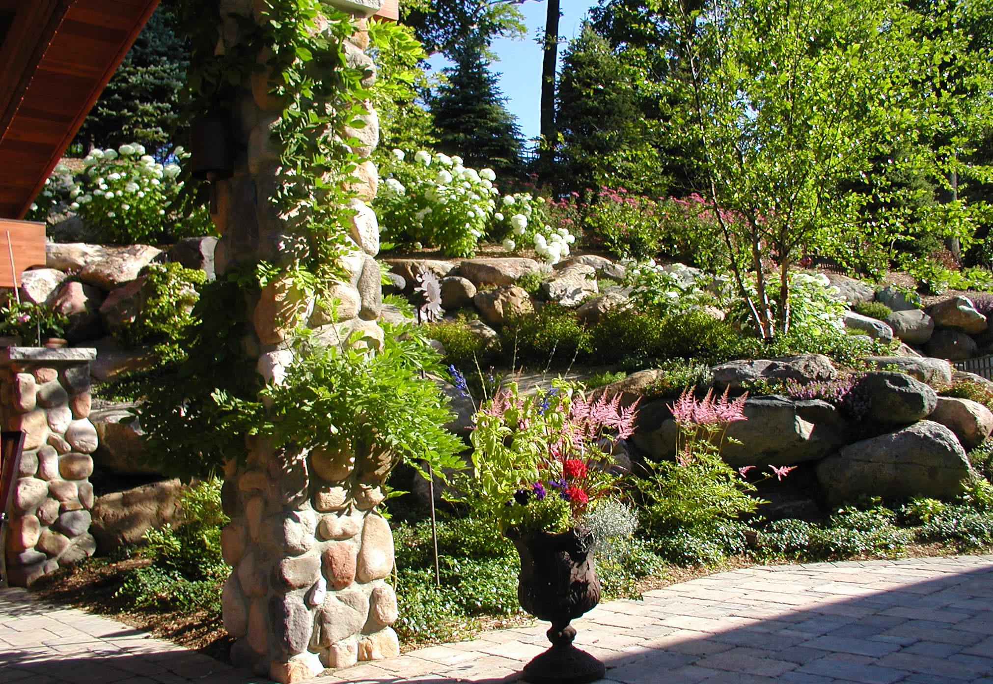 Stone patio with a vine-covered stone pillar and decorative planter filled with colorful flowers, set against terraced landscaping with boulders, hydrangeas, and small trees.
