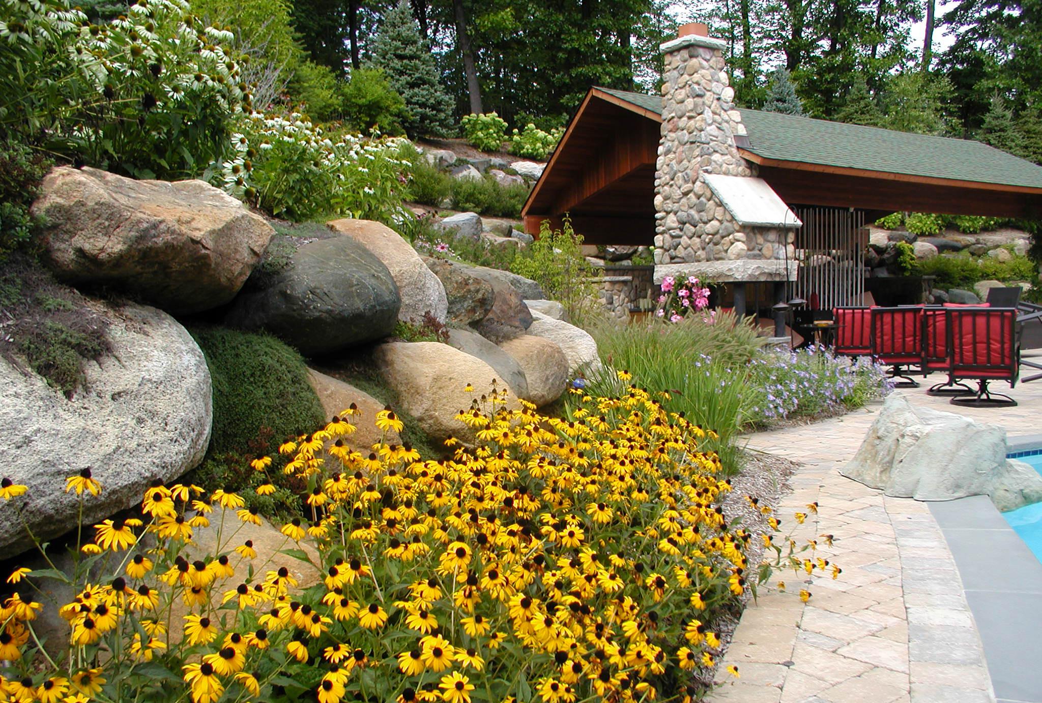 Stone patio with yellow black-eyed Susan flowers in the foreground, large boulders on a hillside, and an outdoor stone fireplace with red cushioned chairs near a swimming pool.