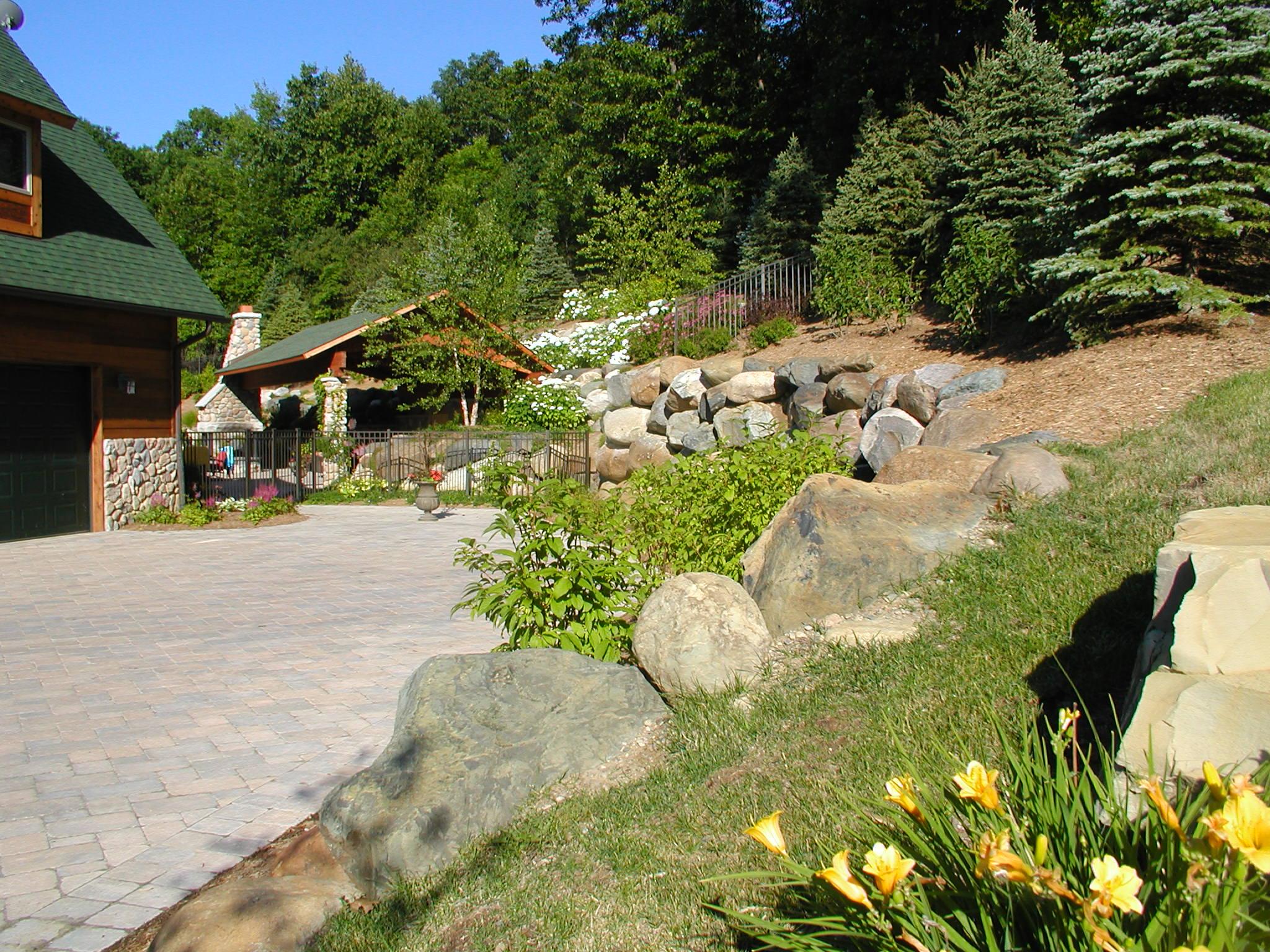Stone-paved driveway bordered by large boulders and landscaped greenery, with a log-style house and outdoor stone fireplace pavilion in the background.