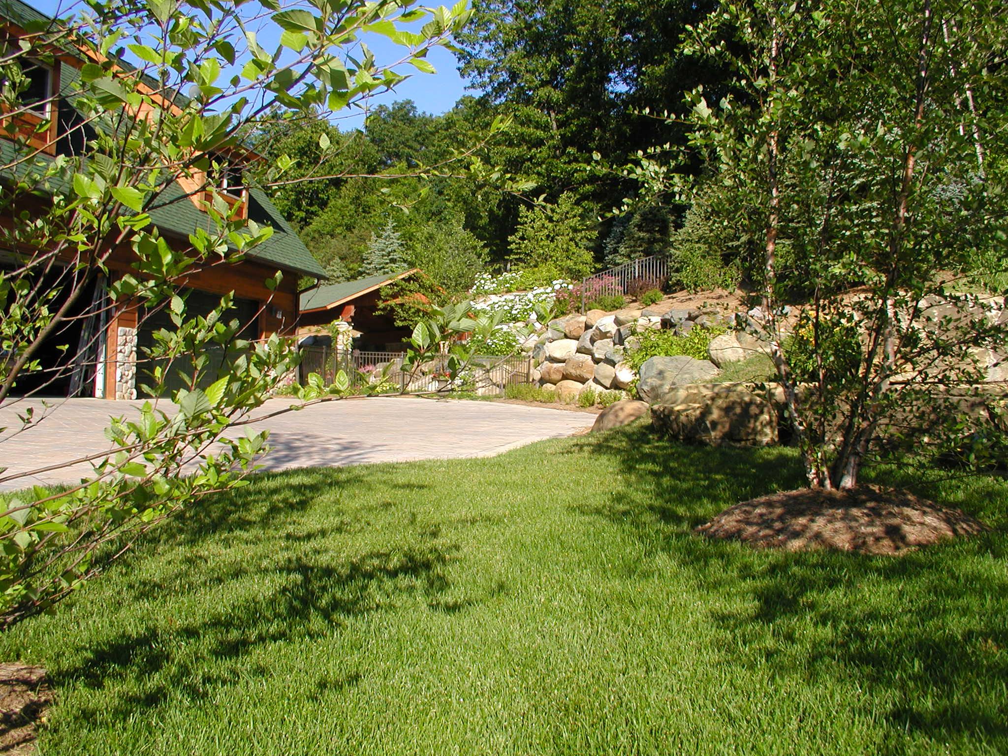 View of a green lawn with young trees in front of a house with green roof and stone details, landscaped with boulders and flowering plants on the hillside.